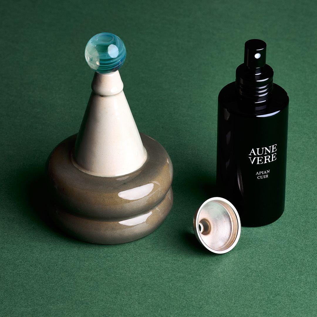 Apian Cuir ceramic vessel with its 100 ml bottle and a silver funnel, assembled on green paper.
