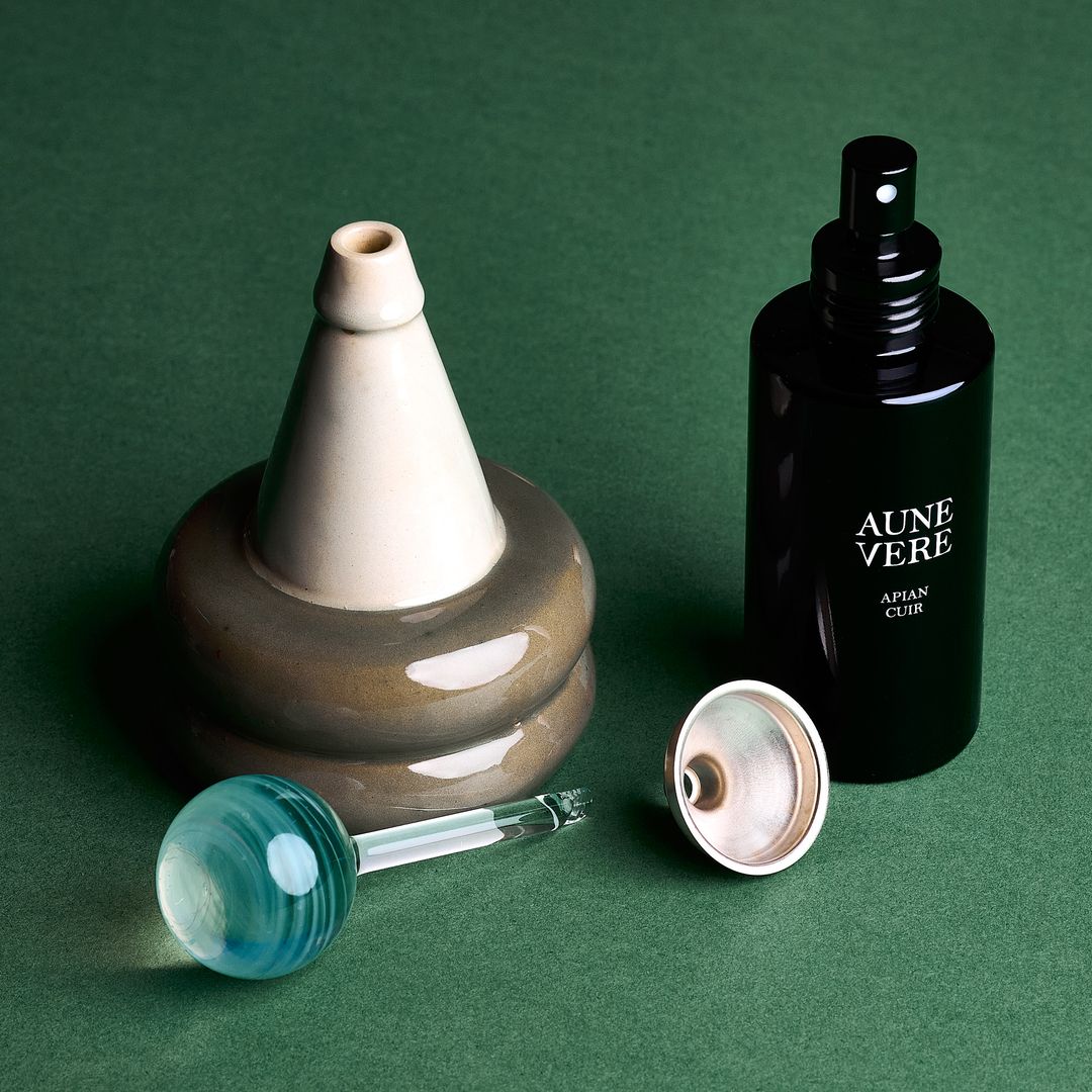 Apian Cuir ceramic vessel open beside its teal glass applicator, 100 ml bottle, and a silver funnel, on green paper.