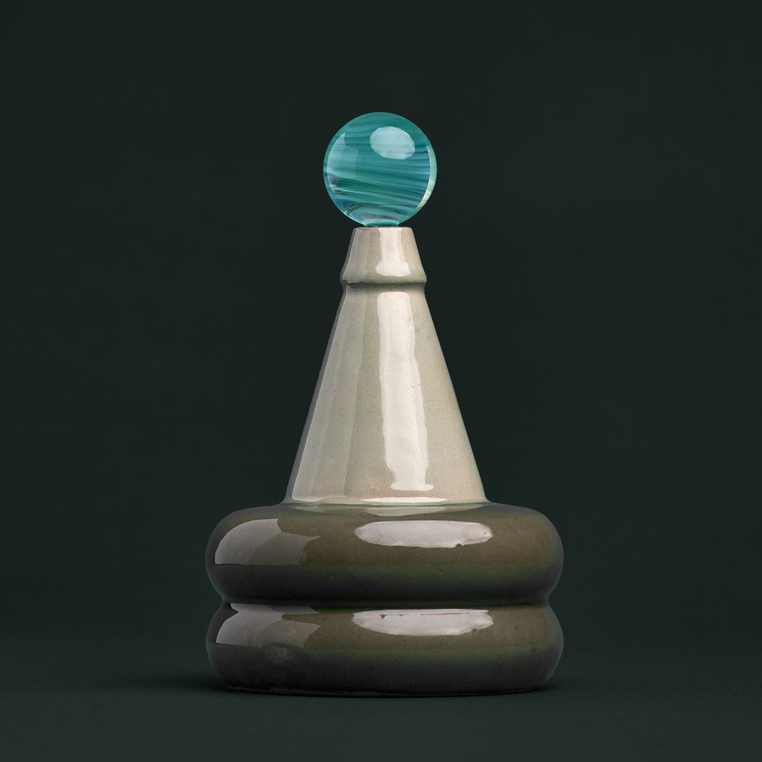 Apian Cuir ceramic vessel, a stacked glazed form holding a teal glass orb, on gray green paper.
