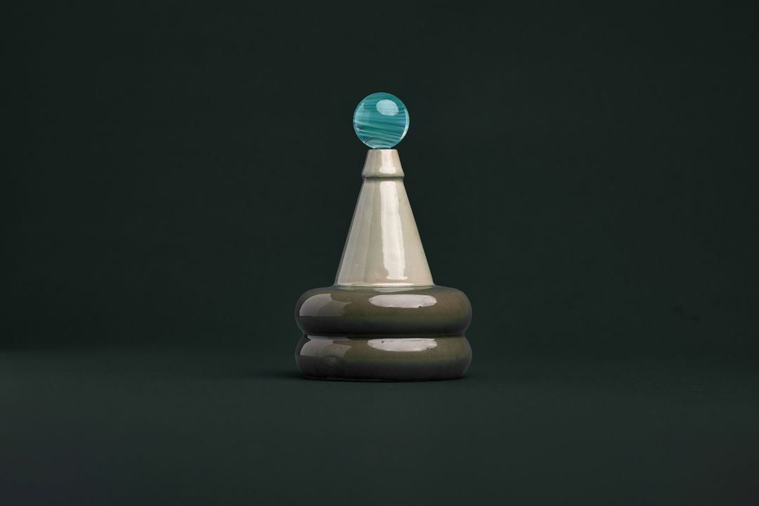 Apian Cuir ceramic vessel, a faceted dark glazed base holding a clear glass orb, on gray green paper.