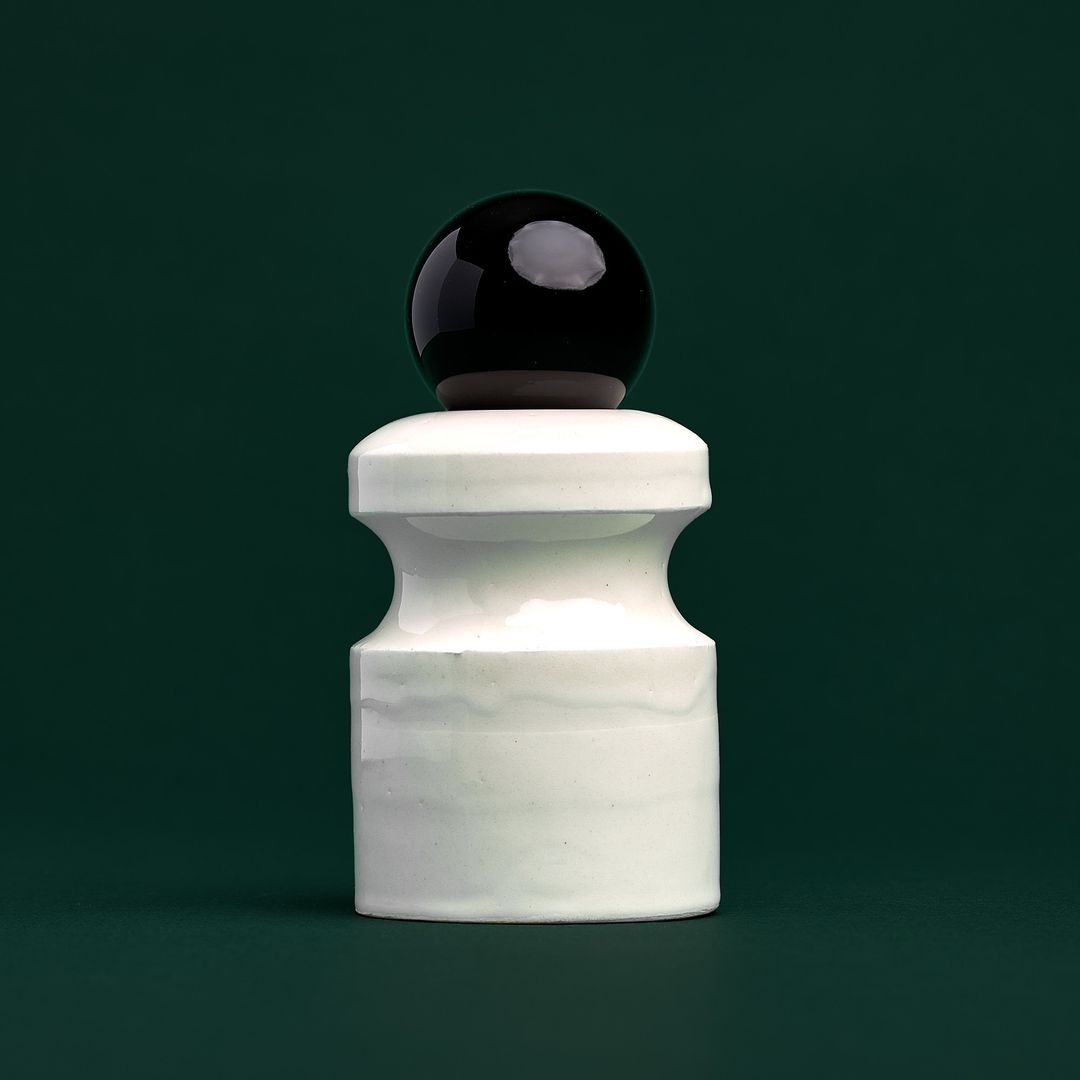 Caledonian Pear ceramic vessel, a pale glazed form holding a dark glass orb, on mossy green paper.
