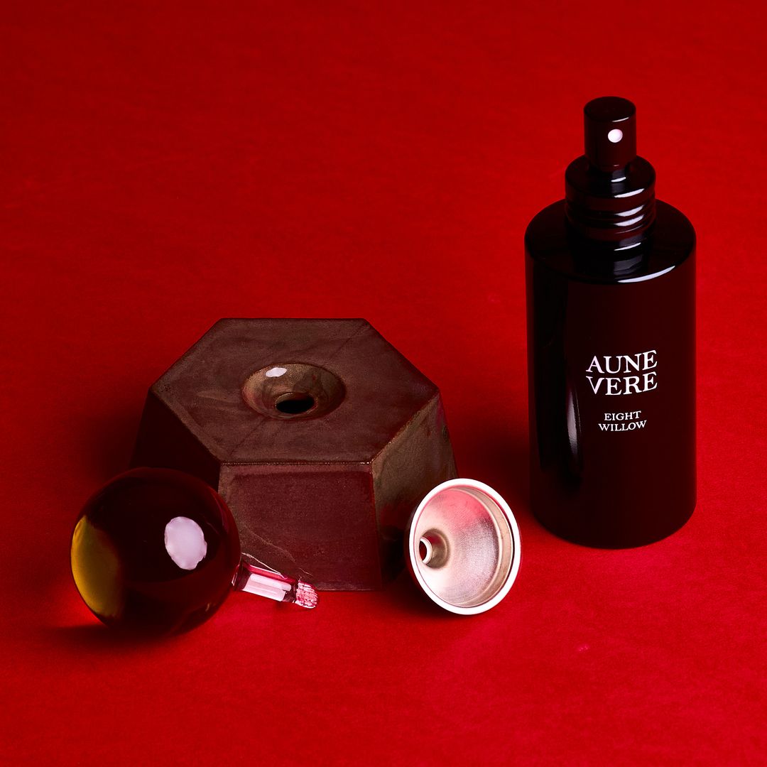Eight Willow ceramic vessel open beside its amber glass applicator, 100 ml bottle, and a silver funnel, on alder red paper.