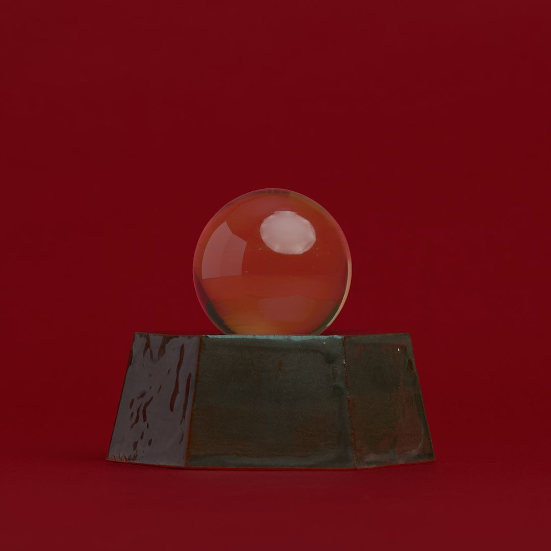 Eight Willow ceramic vessel, a faceted dark glazed base holding an amber glass orb, on alder red paper.