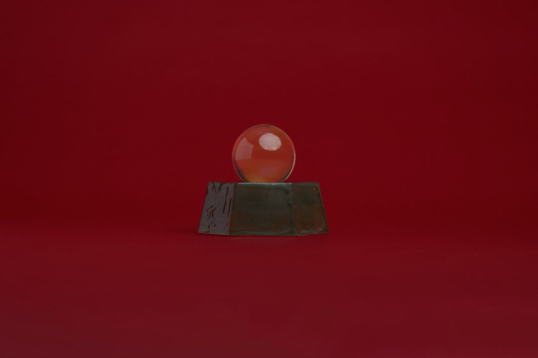 Eight Willow ceramic vessel, a faceted dark glazed base holding a clear glass orb, on alder red paper.