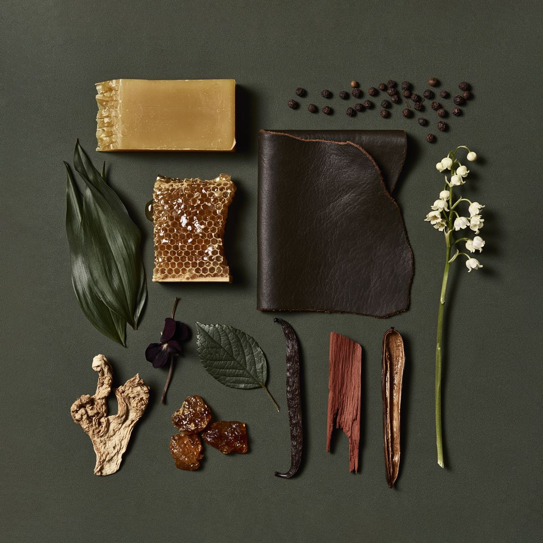 Apian Cuir ingredients laid on green paper: honeycomb, beeswax, leather, pepper, vanilla, woods, and a spray of white flowers.