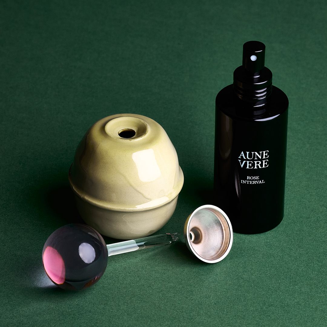 Rose Interval ceramic vessel open beside its pink glass applicator, 100 ml bottle, and a silver funnel, on green paper.