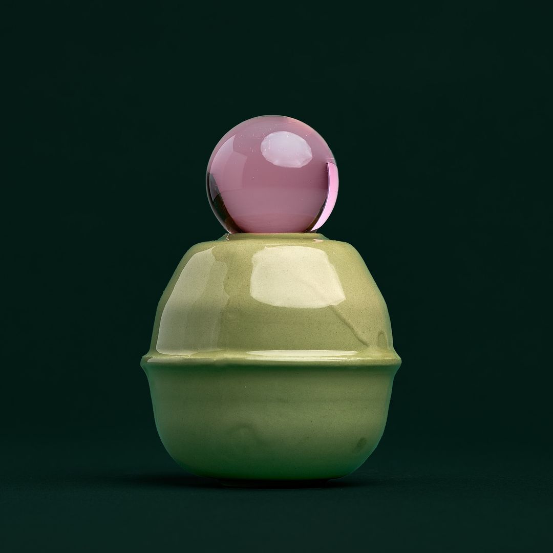 Rose Interval ceramic vessel, a pale green glazed form holding a pink glass orb, on deep bottle green paper.